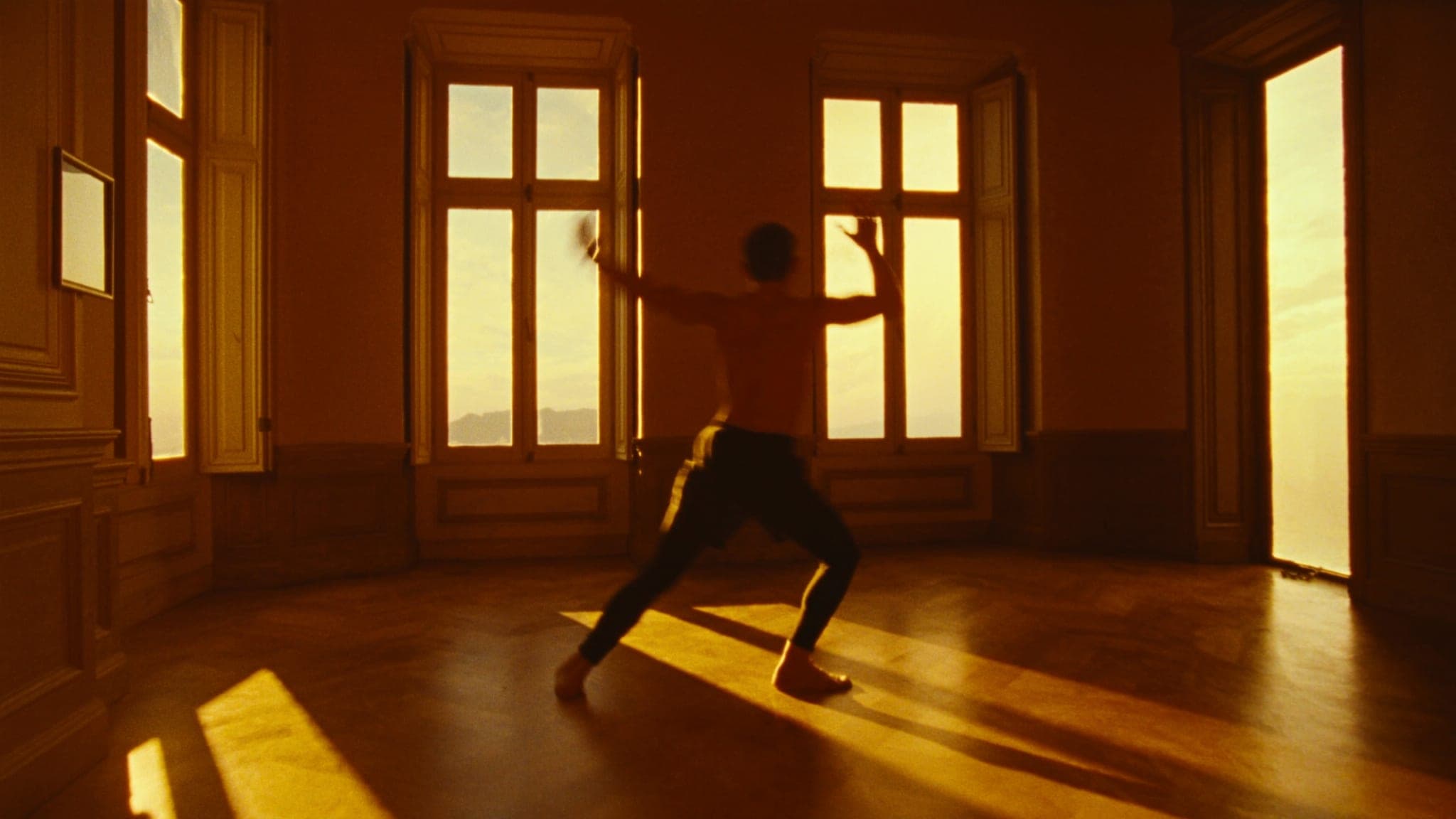 Il salone di una villa al tramonto, luce d’oro dalle finestre alte e una persona in affondo con le braccia aperte.