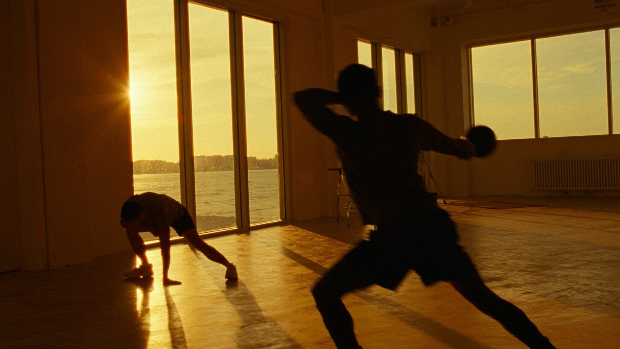 Uno studio al tramonto, luce radente sul pavimento e due corpi in movimento controluce.