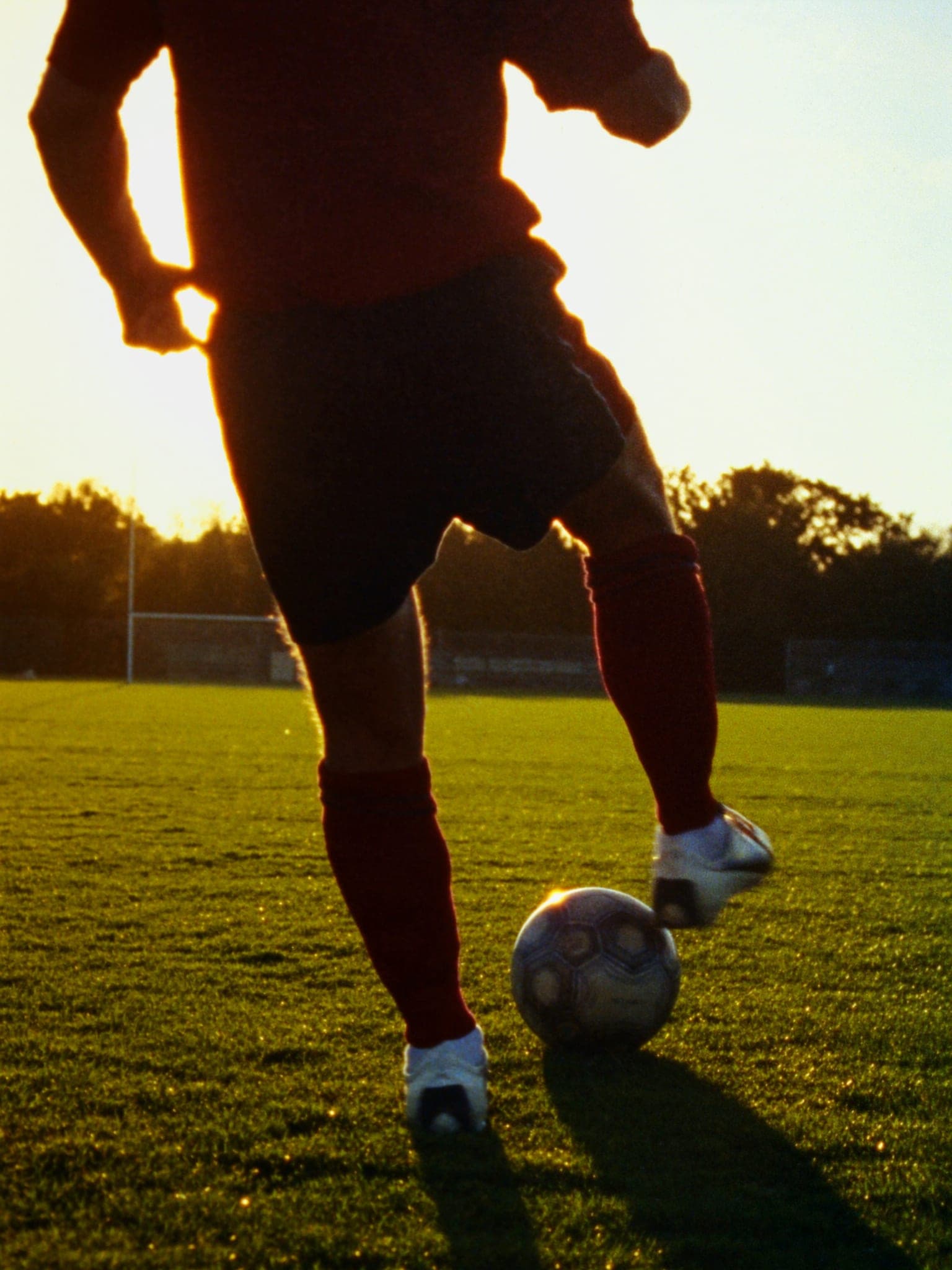 Un calciatore di spalle che calcia di collo, gamba d’appoggio piantata, sole basso sull’erba.
