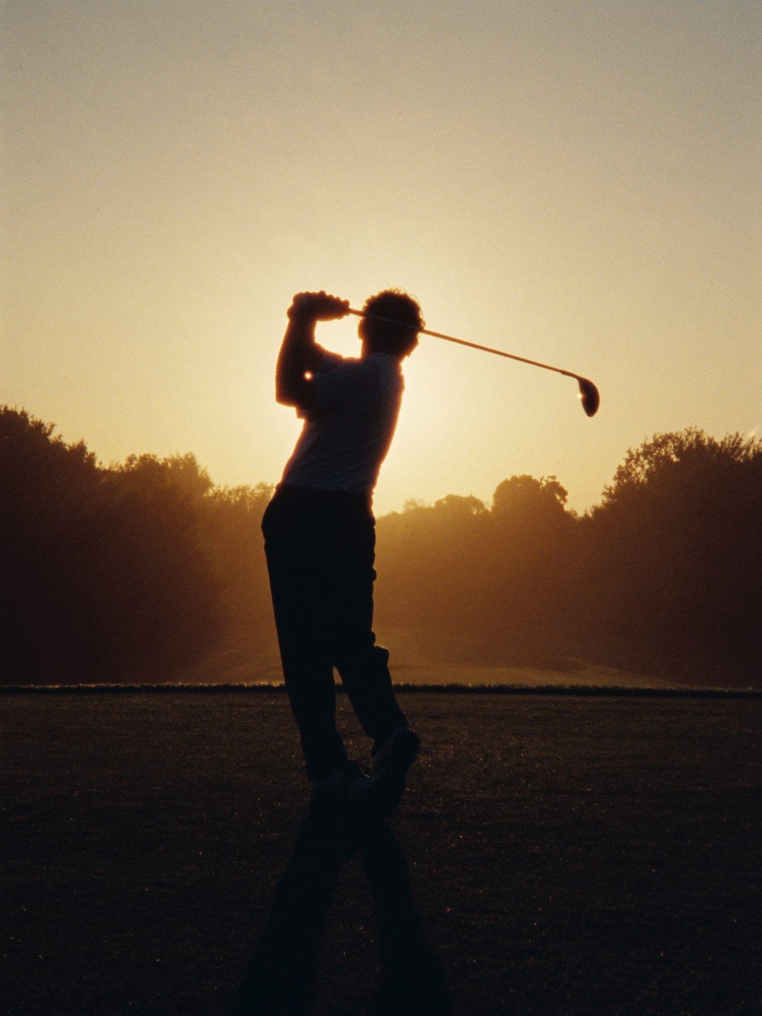 Un golfista di spalle in cima al backswing, controluce al tramonto sul fairway.