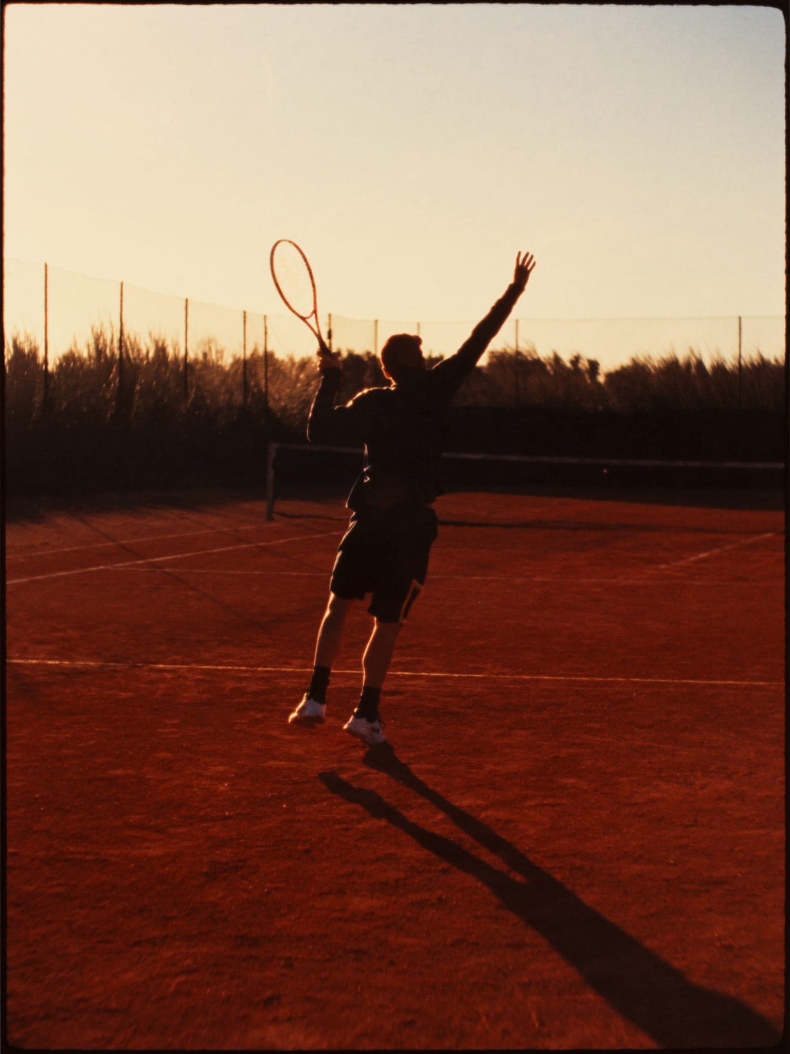 Un tennista di spalle nel servizio, corpo disteso, luce radente sulla terra rossa.