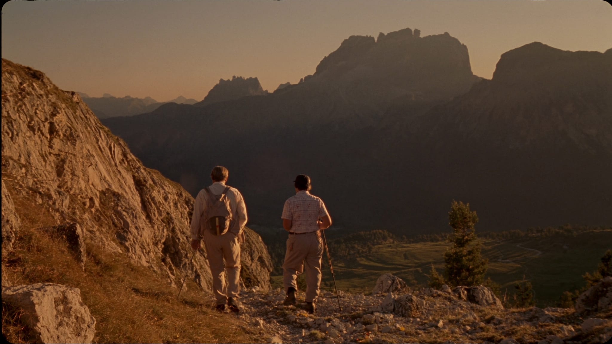 Due persone di spalle camminano su un sentiero di roccia nelle Dolomiti, al tramonto: una con lo zaino, l’altra con il bastoncino, le cime in controluce davanti a loro.