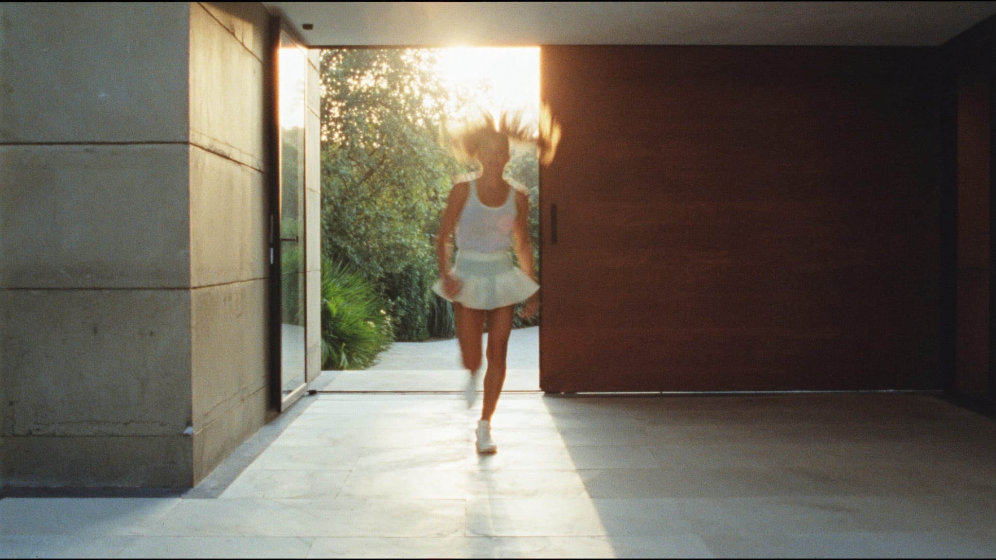 Il portone di una villa moderna aperto sul giardino al tramonto; una ragazza in gonna da tennis entra di corsa, coda al vento, volto mosso.